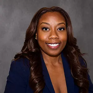 Professional headshot of a smiling woman with long dark hair in a navy blazer against a gray studio backdrop.